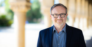 Aron Rodrigue wearing glasses, a blue collared shirt, and a navy-blue blazer standing in one of the arcades on Stanford's Main Quad