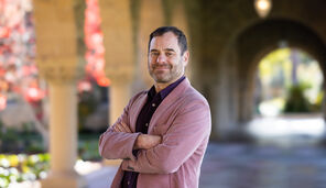 Richard Meyer stands with his arms crossed wearing a mauve blazer over a dark purple shirt.