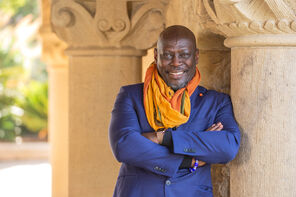 Ato Quayson, wearing a royal-blue blazer and a yellow-gold scarf, leans against a pillar