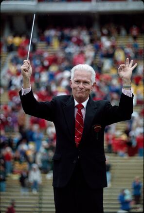 Arthur Barnes conducting the Leland Stanford Junior University Marching Band in the stadium at Stanford. He holds a baton in his right hand and is making an "okay" sign with his left hand while smiling to the band.
