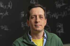Brian Conrad in front of a blackboard filled with math figures wearing a kelly-green sweater over a light-blue collared shirt and a bright-yellow T-shirt