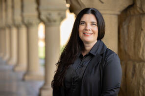 Photo of Michelle Davis with long dark hair wearing a black blouse and standing in one of the arcades with sandstone arches and pillars near Stanford's Main Quad