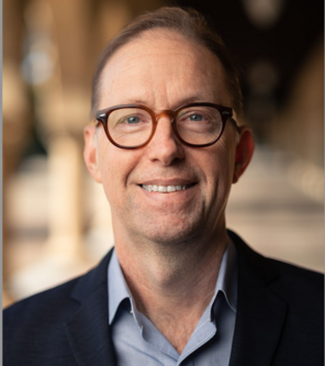 Russell Poldrack wearing glasses, a light-blue collared shirt, and a navy blazer standing in one of the arcades along Stanford's Main Quad