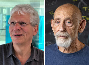 Physicists Stephen Shenker with white hair and wearing glasses and a polo shirt and Leonard Susskind with a short white beard, wearing a crew-neck blue shirt and standing in front of a blackboard full of equations
