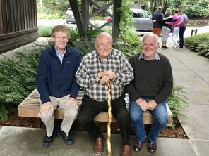 Three older men sit on a bench in front of a some ferns in a courtyard. They are smiling for the photo.
