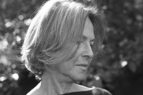 Poet Louise Glück in a black-and-white image. She is looking down and away from the camera. Her chin-length hair is obscuring her right cheek and eye. Sunlight is illuminating the back and crown of her head.