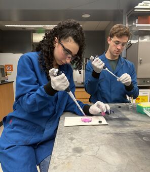 Two students in blue lab coats use pipettes in a lab