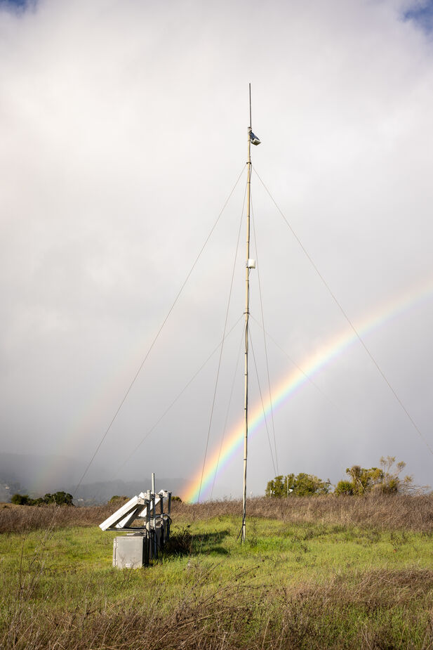 A double rainbow, with the top arc much lighter than the bottom, is seen on top of gray clouds behind a radio antenna and other equipment on top of a grassy hill