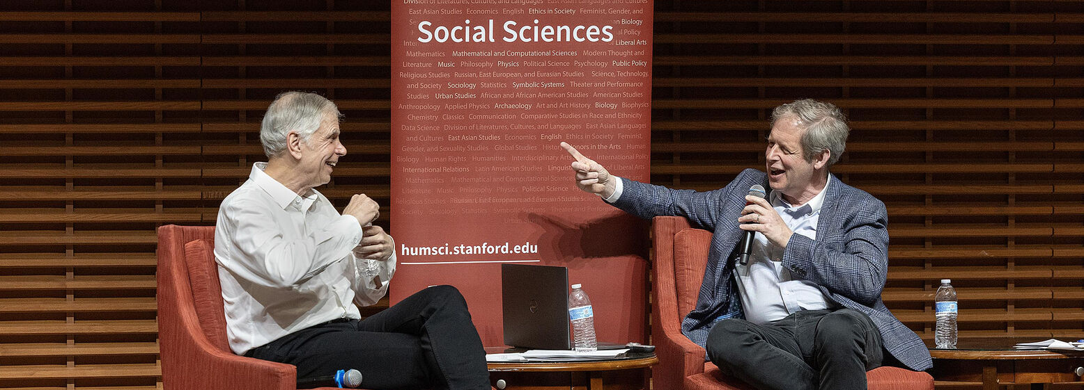 Professors John Donohue and Eugene Volokh wearing business attire, sitting in red chairs on a stage, and using their hands as part of an animated discussion