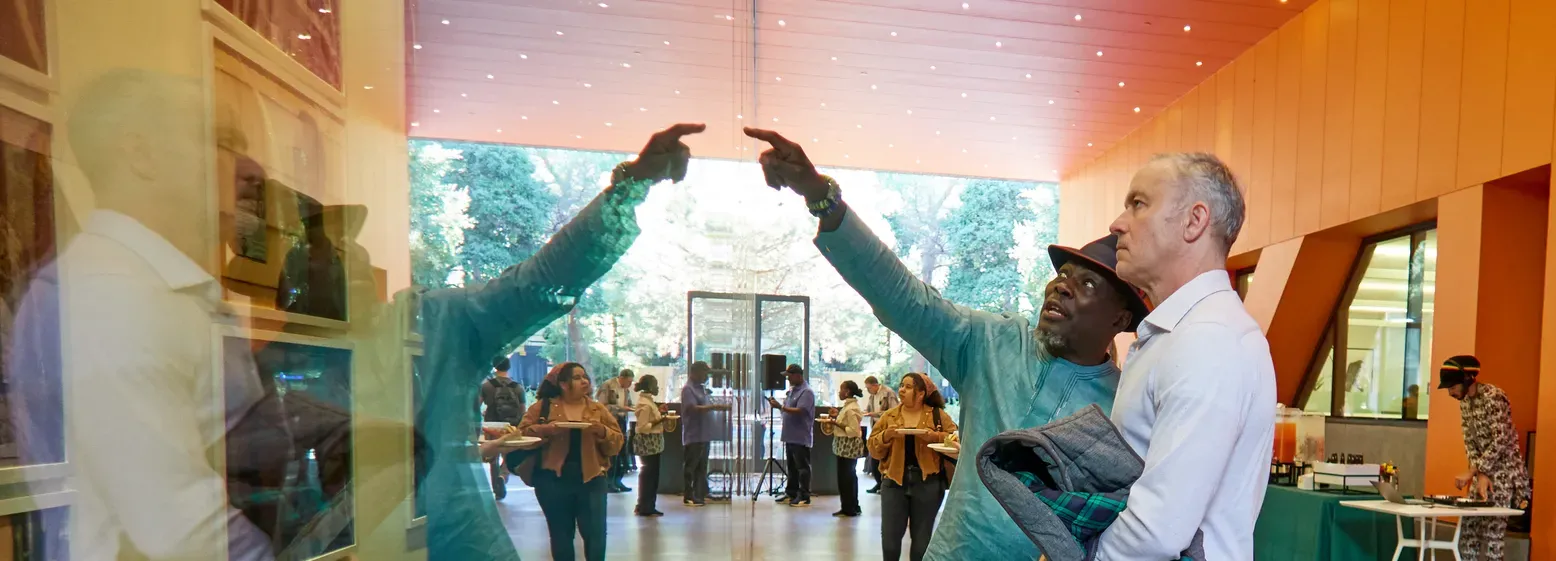 One man points at photos behind glass while another man looks intently. The glass reflects the men's images.