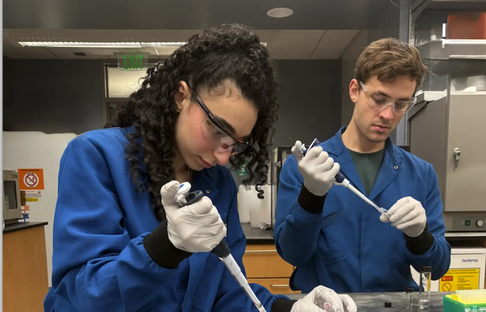 A female and a male scientist wearing gloves, safety glasses, and blue lab coats work with large pipettes at a lab table