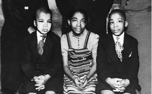 A black-and-white photo of three young black children sitting on a bench