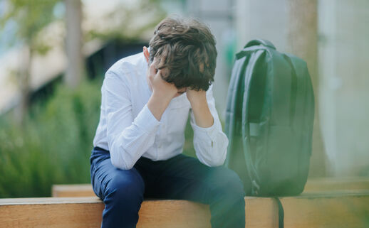 A photo of a young school boy sitting on a bench with his head in his hands.