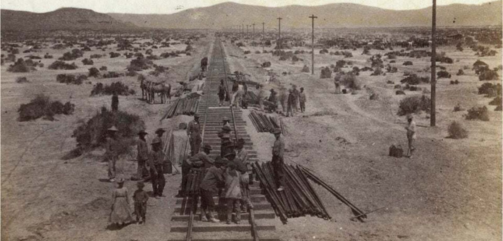 Laborers lay railroad track through a desert with mountains on the horizon in an 1860s black-and-white image