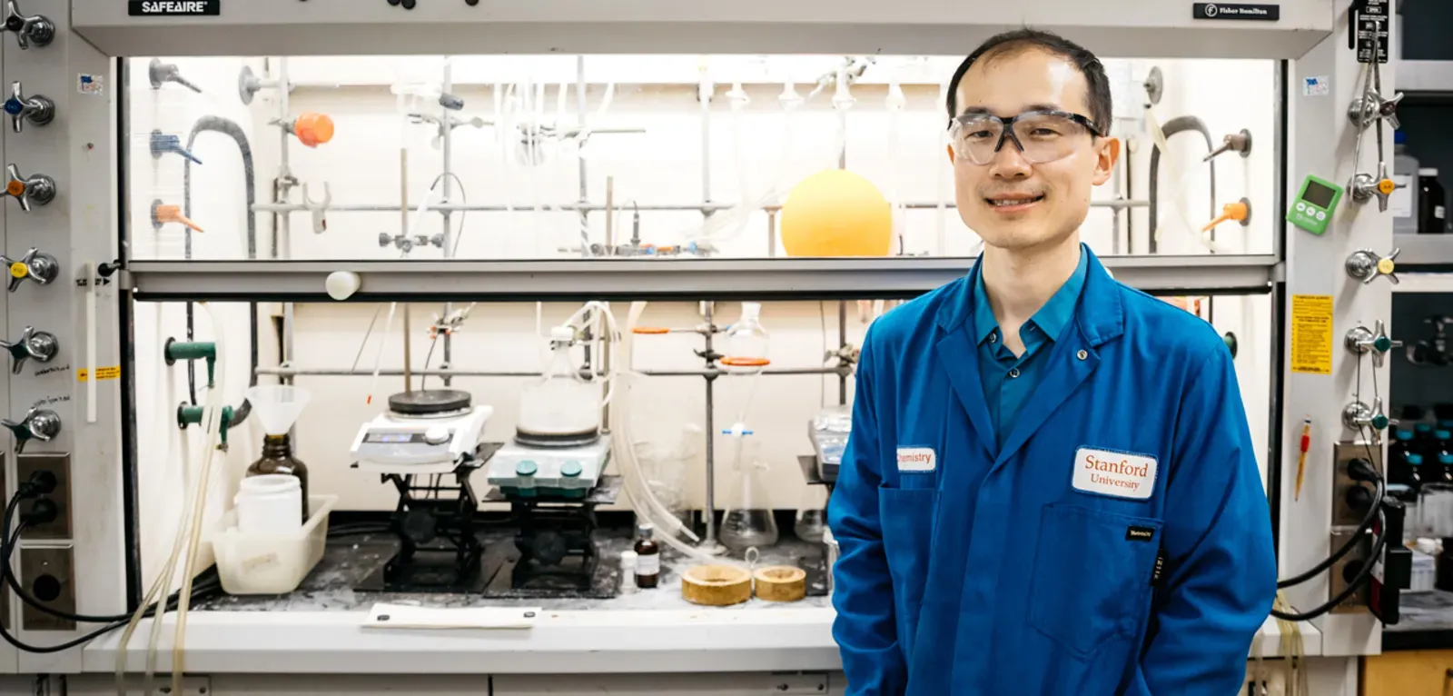 Song Lin stands in his lab wearing safety goggles and a blue lab coat
