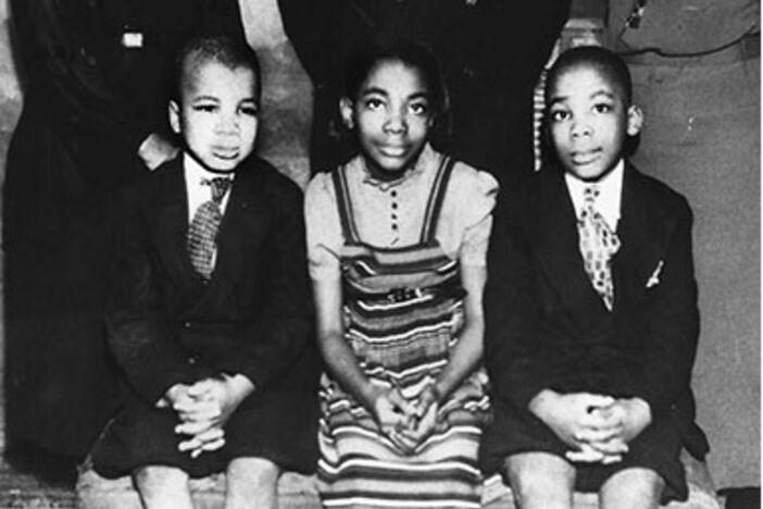 A black-and-white photo of three young black children sitting on a bench