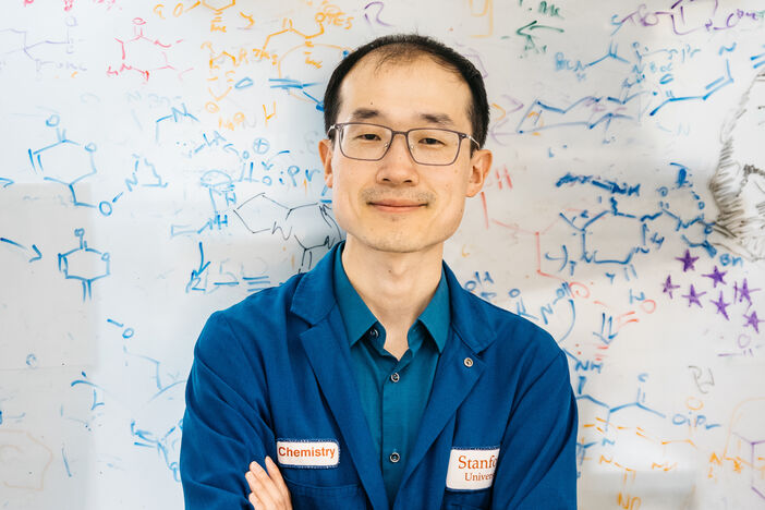 Song Ling wears a blue lab coat and stands in front of a whiteboard filled with drawings of organic molecules