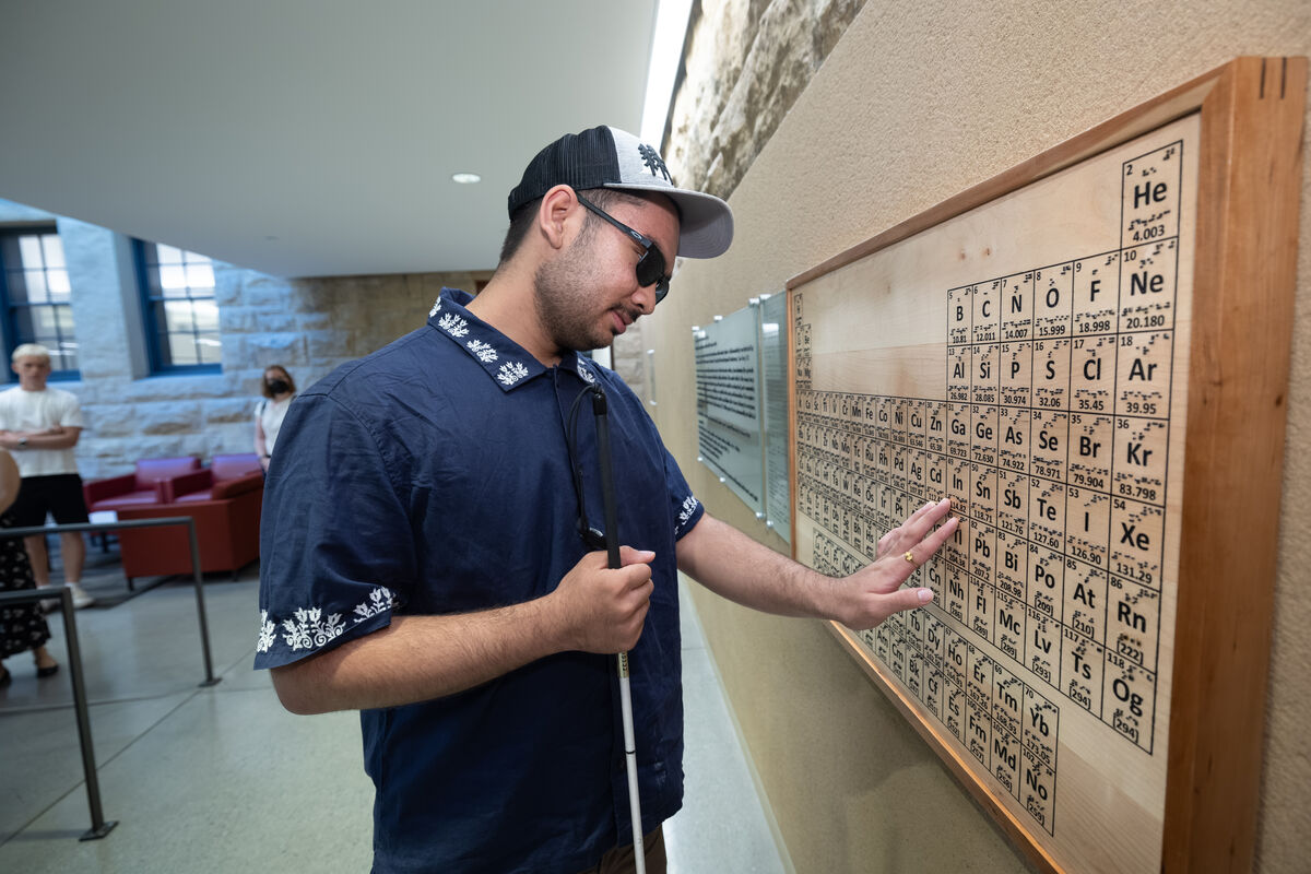 A man holding a cane touches a wooden periodic table of elements mounted on a wall.