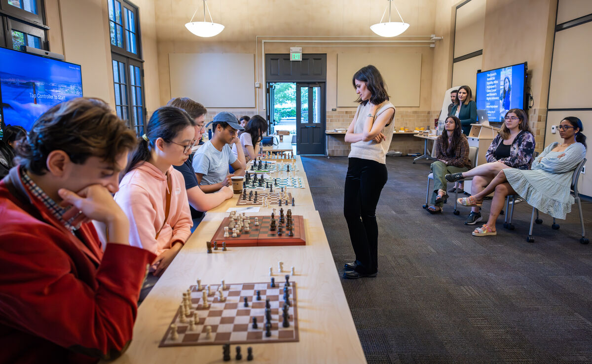 A woman stands in front of a long table with six chess boards and six seated players