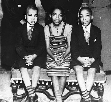 A black-and-white photo of three young black children sitting on a bench