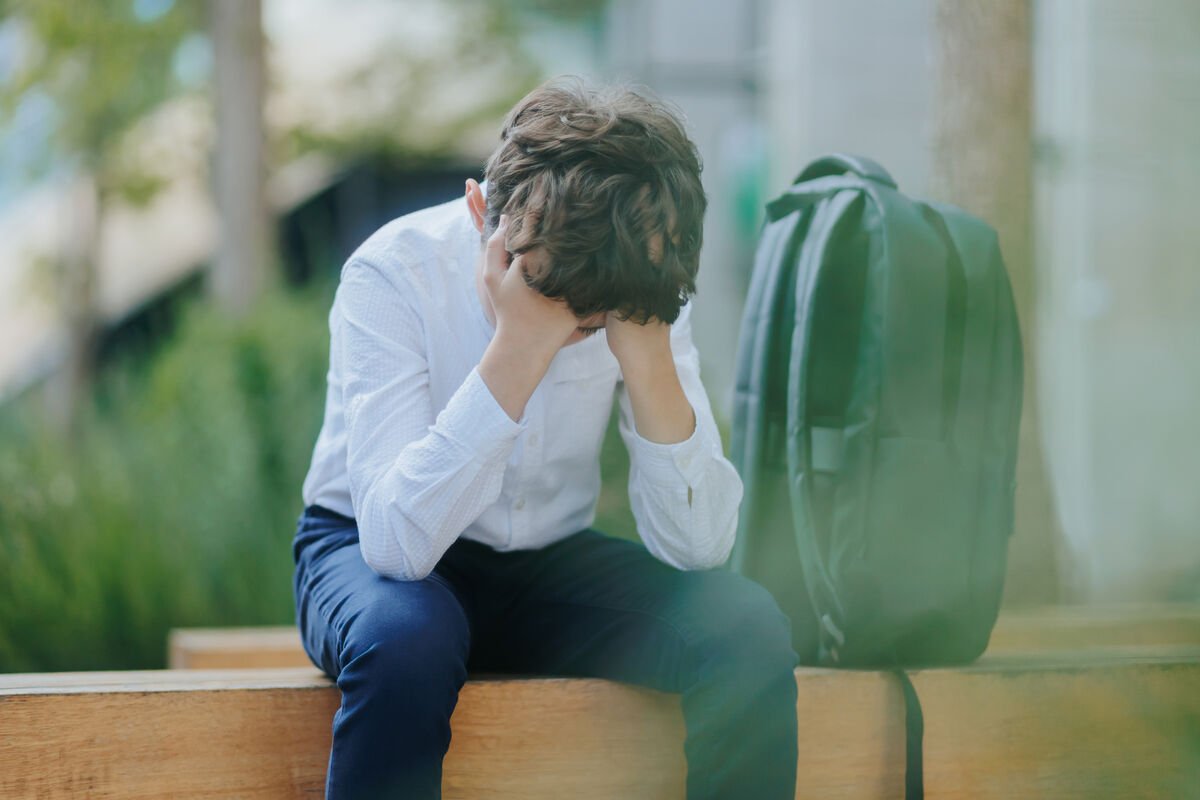 A photo of a young school boy sitting on a bench with his head in his hands.