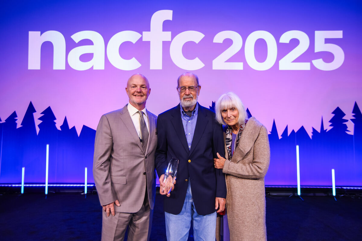 Three people in formal clothes pose for a portrait at an awards the ceremony. Jeff Wine is in the middle holding an award trophy.