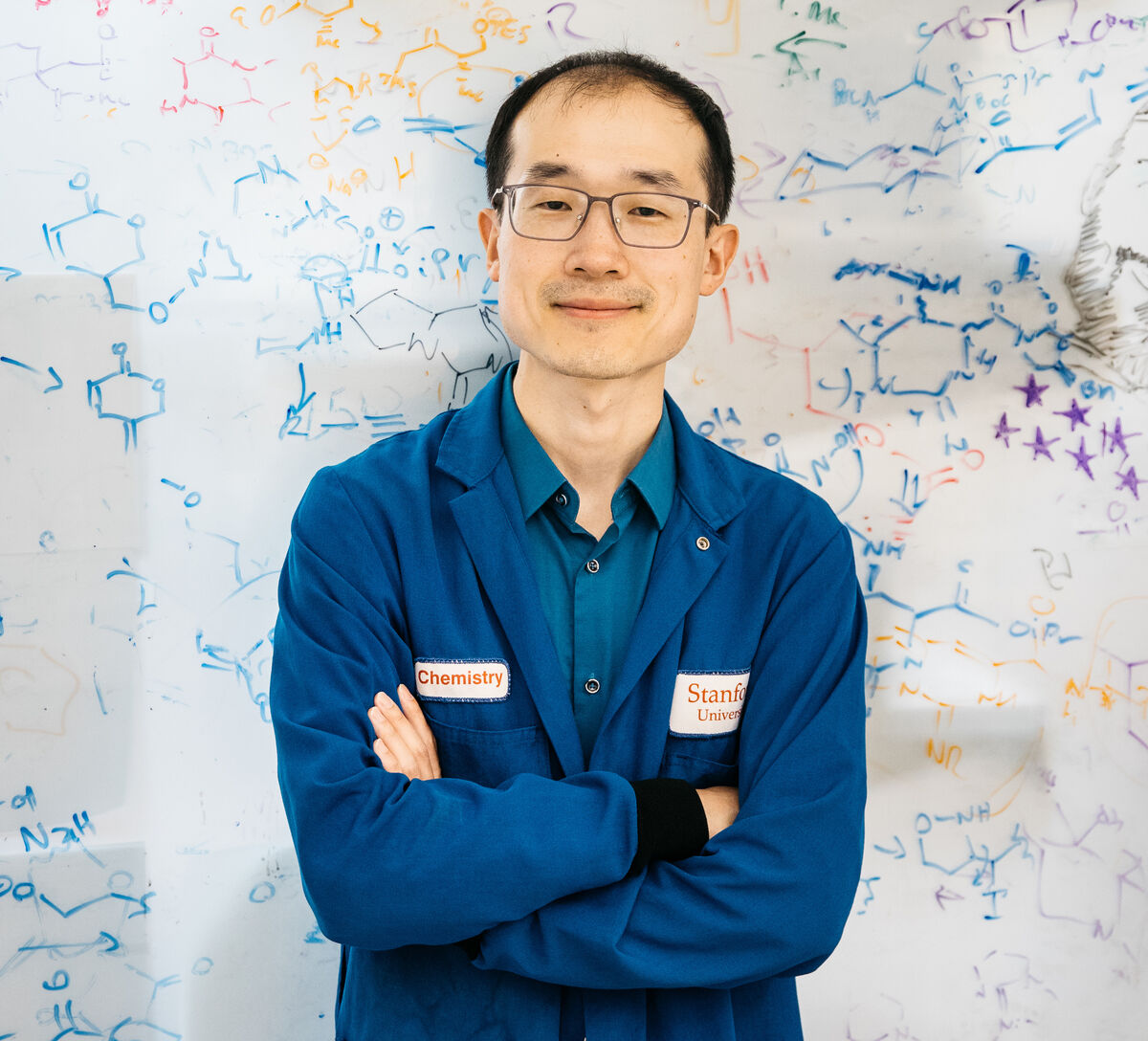 Song Ling wears a blue lab coat and stands in front of a whiteboard filled with drawings of organic molecules