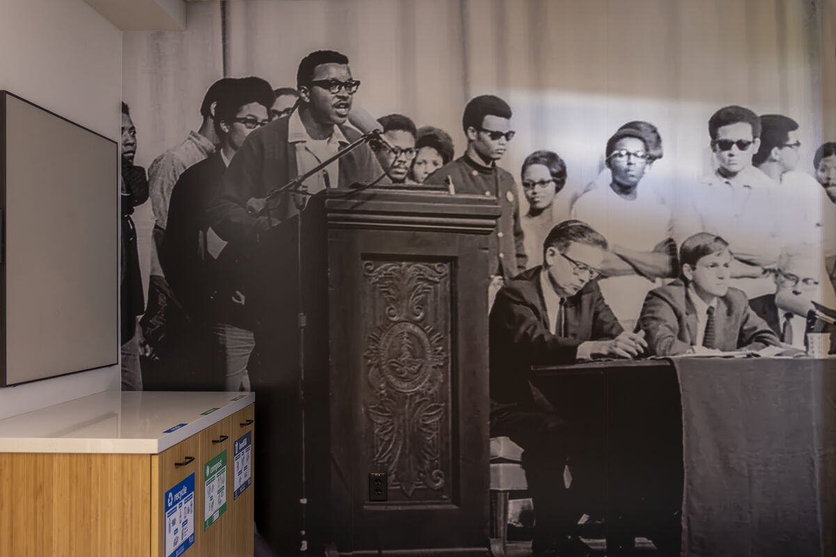 A wall-sized black-and-white photo shows a Black man standing at a podium surrounded by other Black men next to White men sitting a a table.