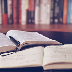 Two books lie open on a table, one with a pen in the centerfold. There is a shelf filled with books in the background.