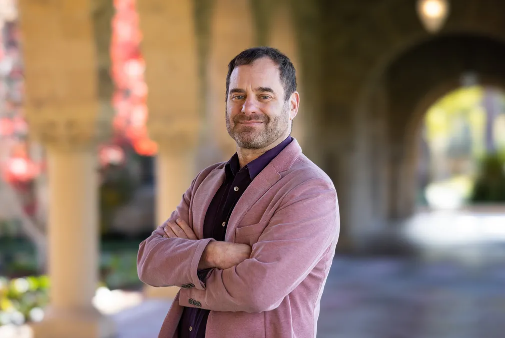 Richard Meyer stands with his arms crossed wearing a mauve blazer over a dark purple shirt.