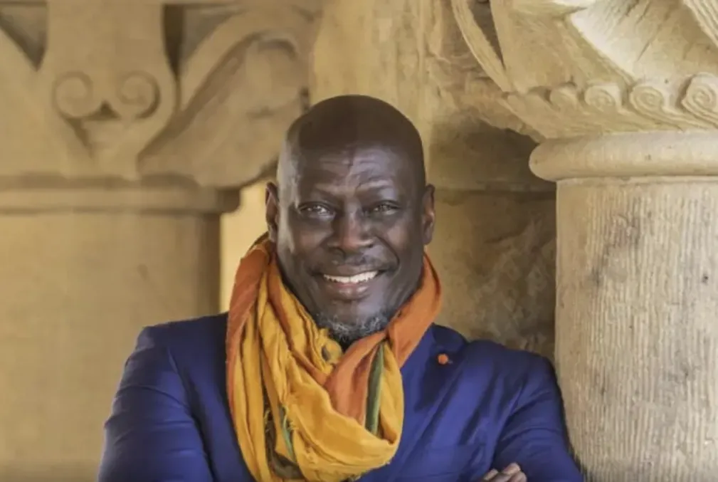 Ato Quayson, wearing a royal-blue blazer and a yellow-gold scarf, leans against a pillar