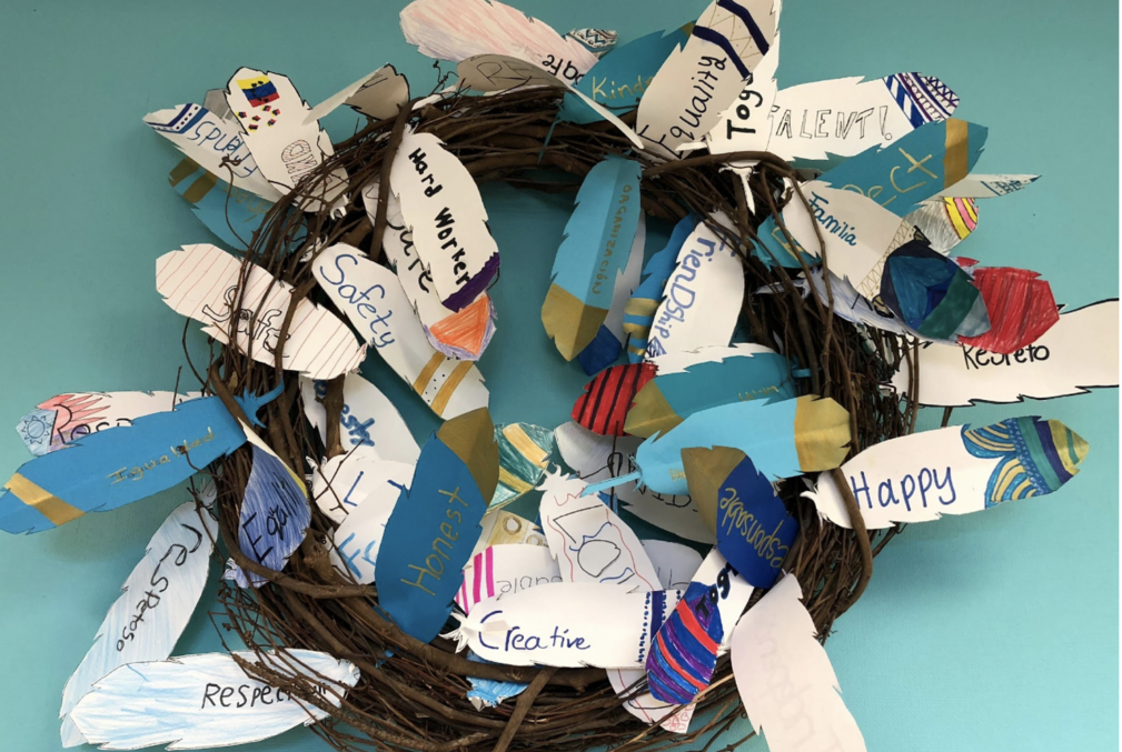Image of a wreath with paper feathers on it. The paper feathers have writing on them that represent the values that each student's family cherishes.