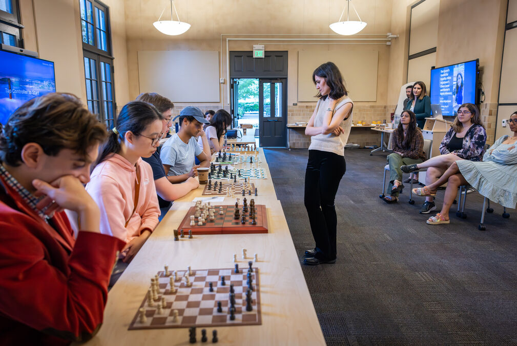 A woman stands in front of a long table with six chess boards and six seated players