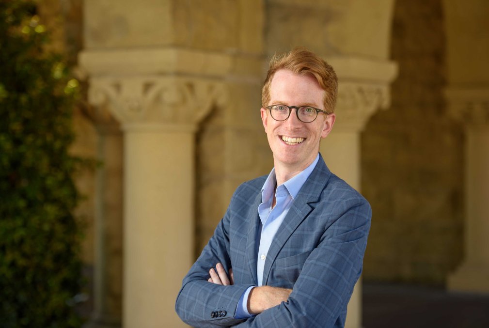 Rowan Dorin wearing glasses, a light-blue collared shirt, and a medium-blue blazer standing with arms crossed in front of columns along Stanford's Main Quad