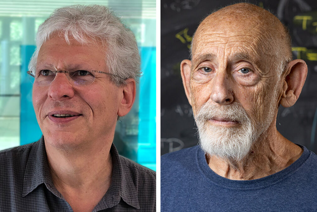 Physicists Stephen Shenker with white hair and wearing glasses and a polo shirt and Leonard Susskind with a short white beard, wearing a crew-neck blue shirt and standing in front of a blackboard full of equations