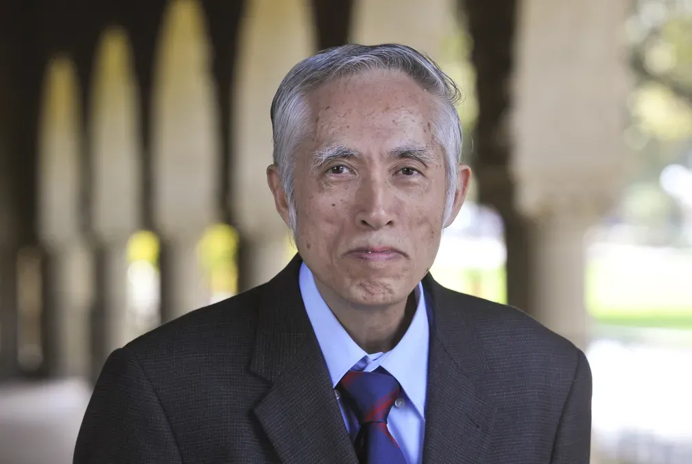 Tze Leung Lai wearing a light-blue collared shirt, royal blue and red tie, and dark coat standing under one of the arcades along Stanford University's Main Quad