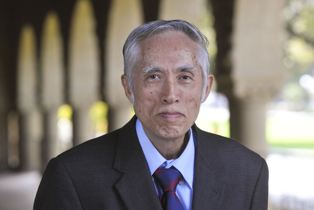 Tze Leung Lai wearing a light-blue collared shirt, royal blue and red tie, and dark coat standing under one of the arcades along Stanford University's Main Quad