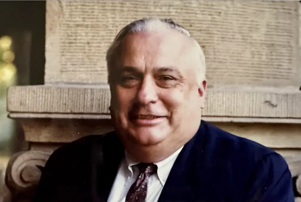 Walter Falcon wearing a navy-blue coat, white collared shirt, and a dark tie in front of a sandstone pillar 
