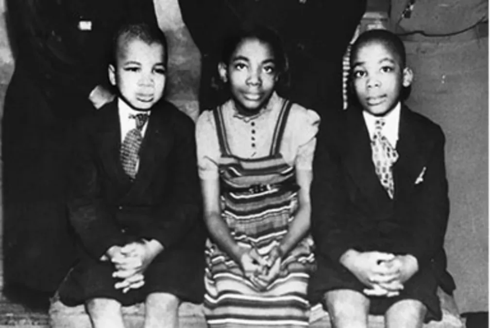 A black-and-white photo of three young black children sitting on a bench