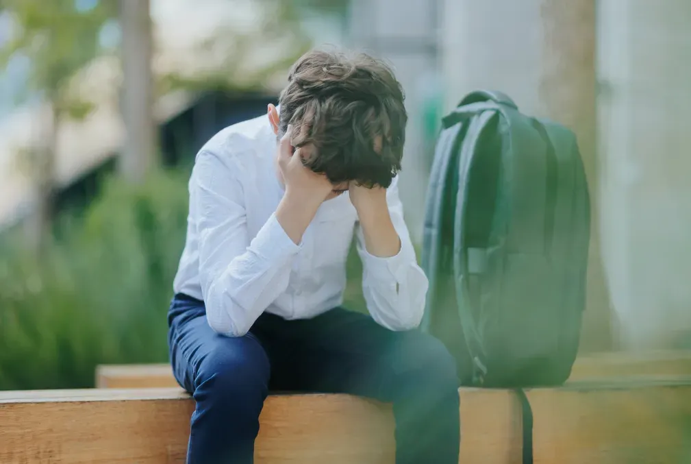 A photo of a young school boy sitting on a bench with his head in his hands.