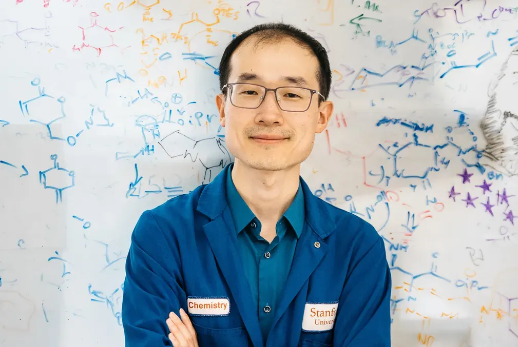 Song Ling wears a blue lab coat and stands in front of a whiteboard filled with drawings of organic molecules