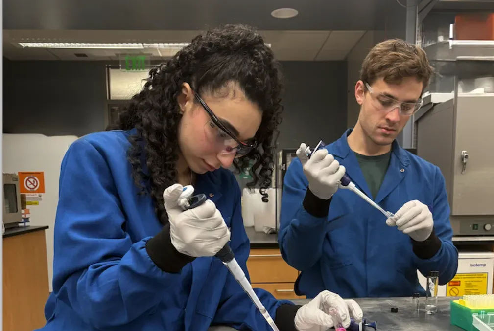 A female and a male scientist wearing gloves, safety glasses, and blue lab coats work with large pipettes at a lab table