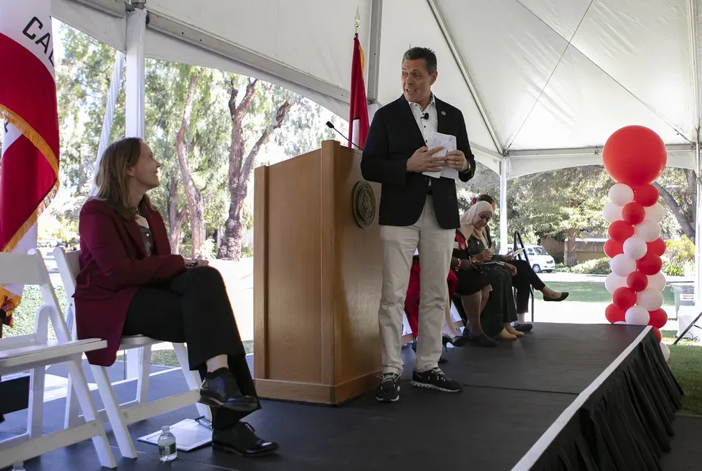 On a stage, a woman sits in a chair and a man stands in front of a lecturn